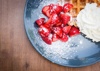Close up of strawberries covered in powdered sugar with Belgian waffles and whipped cream on a blue plate on a wooden table © Dave
