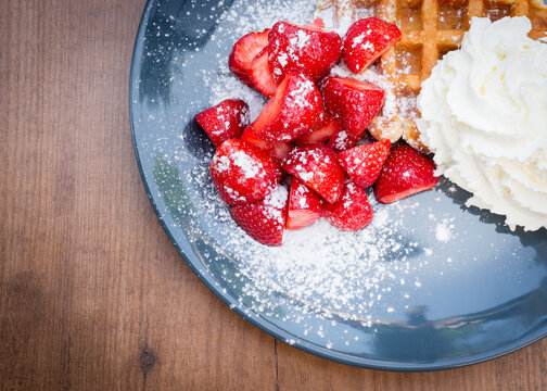 Close up of strawberries covered in powdered sugar with Belgian waffles and whipped cream on a blue plate on a wooden table