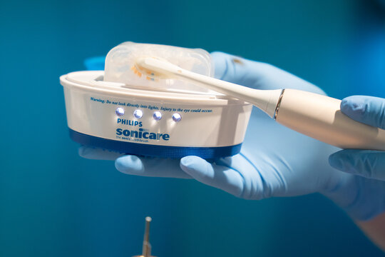 WARSAW, POLAND - FEBRUARY 18, 2026: A close-up view of a person wearing blue medical gloves holding a Philips Sonicare UV toothbrush head sanitizer with a sonic toothbrush placed on top