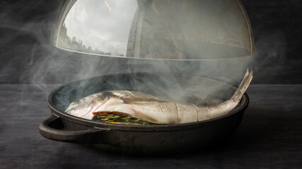 A cooked sea bream with smoke coming from the pan as a glass dome is lifted up in a restaurant © Dave
