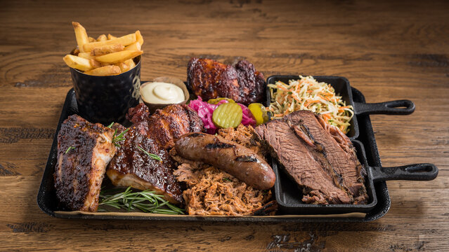 A barbecue meat platter with spare ribs, pulled pork, sausage, beef brisket slices, coleslaw, fires, mayonnaise, chicken pieces and pickles