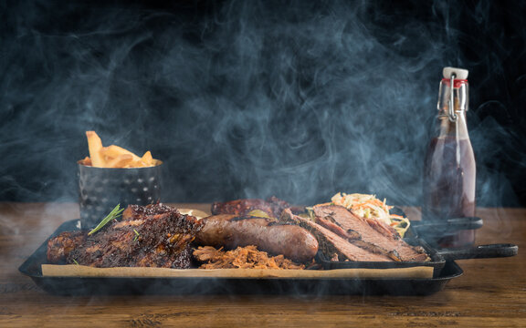 A barbecue meat platter with spare ribs, pulled pork, sausage, beef brisket slices, coleslaw, fires, mayonnaise, chicken pieces, pickles and bbq sauce with smoke surrounding the food