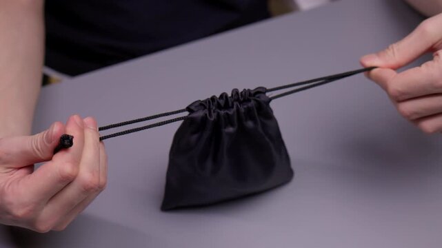 Male hands pulling parallel drawstrings of black fabric storage pouch, gathering top opening to secure contents and achieve organized containment for accessories or gadgets, close-up, slow motion.