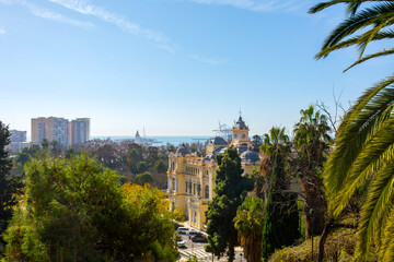 Malaga, Spain &ndash; Malaga City Hall historical architecture with clock tower viewed through lush green park trees in La Malagueta district during a bright day in the coastal city center.