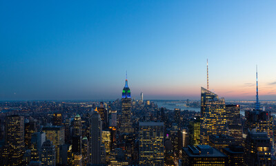 Obraz premium Panoramic view of New York City skyline at dusk featuring illuminated skyscrapers and iconic architecture under a colorful sunset sky. Urban cityscape, travel and business concept.