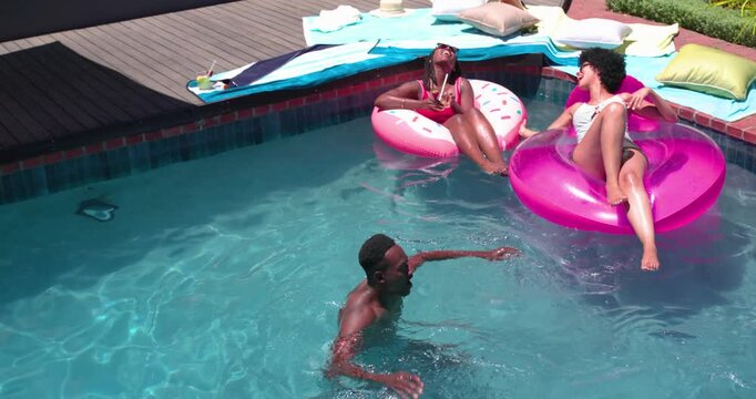African American friends laughing on donut float as man raising arms to play in backyard pool