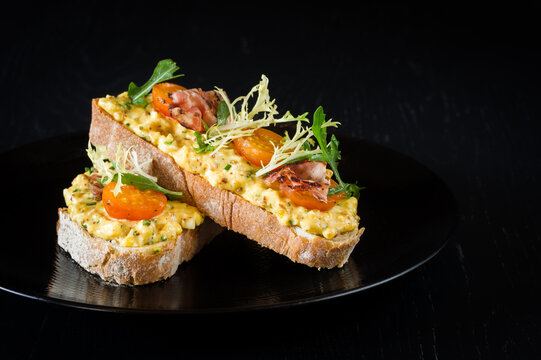 An egg mayonnaise open sandwich with tomatoes, salad and bacon on slices of bread on a plate