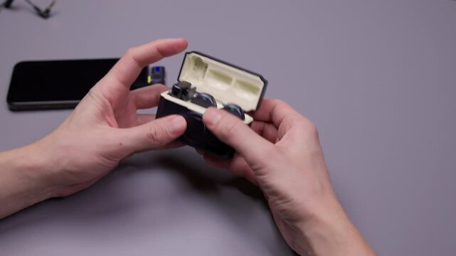 Broadcaster checking compact wireless lapel microphone system for smartphones and vlogging, showing transmitter, receiver, charging case and magnetic clip accessories, close-up, slow motion.