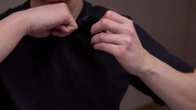 Blogger male puts T-shirt and checks small lavalier microphone with furry windshield, preparing audio equipment for recording, podcasting, video content creation, ensuring clear sound capture.