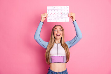 Happy young woman holding a calendar with marked dates on pink background showcasing cheerful and...