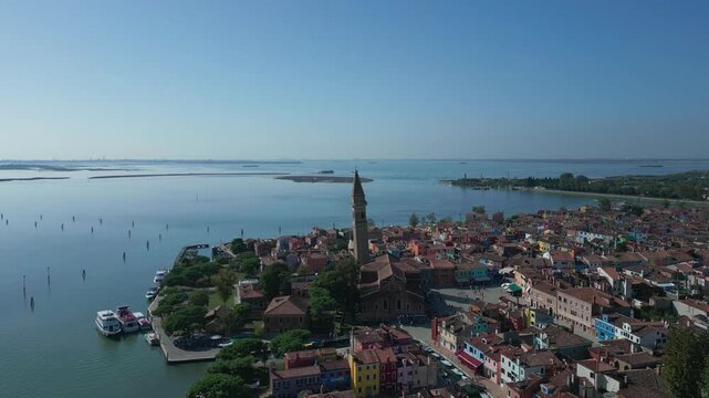 Burano, Venice, Italy: Aerial view of the vibrantly colored houses and bell tower surrounding the lagoon under a clear blue sky.