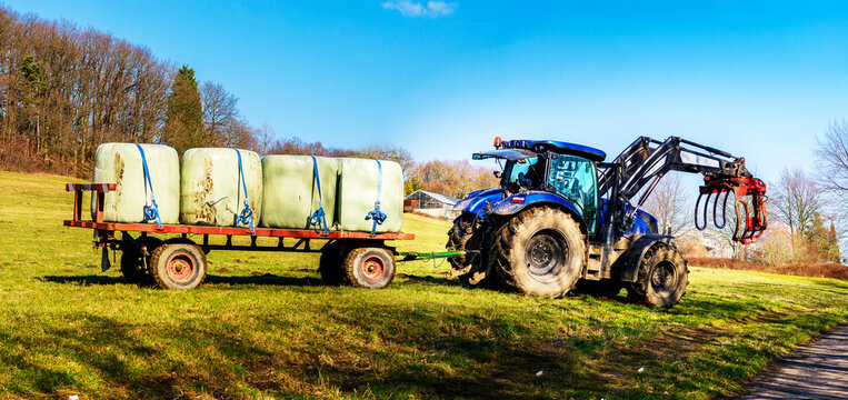 Blue tractor with a trailer full of wrapped hay bales in a sunny field