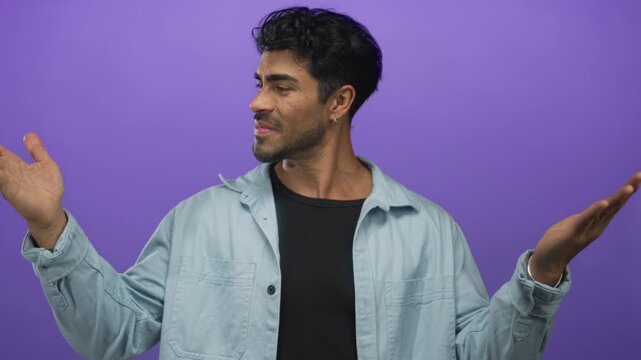 Young hispanic man in a light blue denim shirt holding hands apart measuring width in a purple studio; size evaluation.