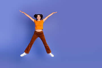Young girl jumps with arms outstretched against violet background wearing orange top brown pants...
