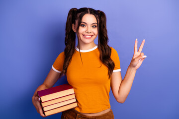 Young female student holds books smiles and shows peace sign against blue background