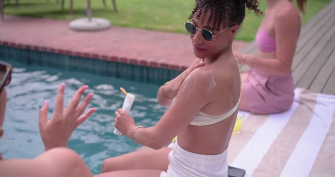 Female friends lounging at private pool, sun warming skin, woman in white bikini applying sunscreen