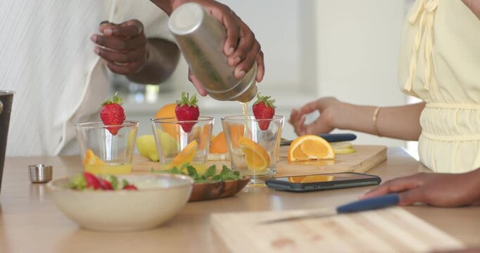 Man bringing shaker, pouring into glasses while woman in yellow dress holding lime making drinks