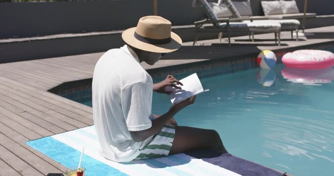 Settling on towel at backyard pool African American man relaxing, turning book pages, drink resting