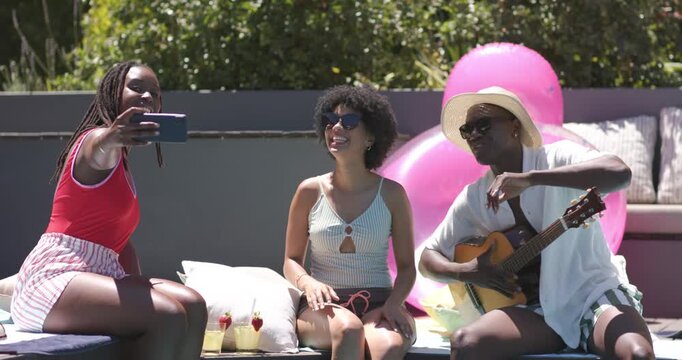 African American friends reacting to phone cue, taking selfies, smiling and playing guitar poolside