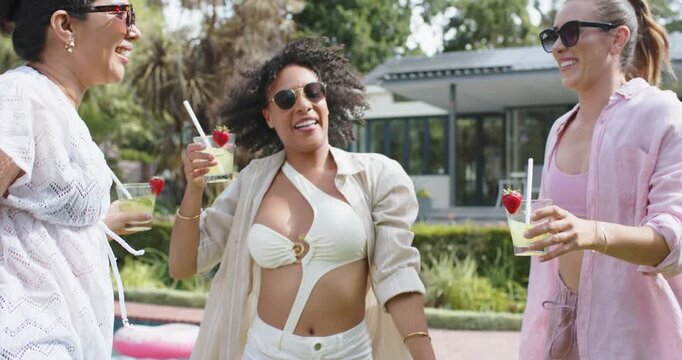 Diverse female friends celebrating walking with central woman lifting arms holding drinks poolside