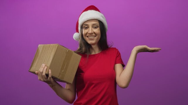 Woman holding cardboard box with palm up gesture in purple studio wearing santa hat and red shirt; holiday cheer.