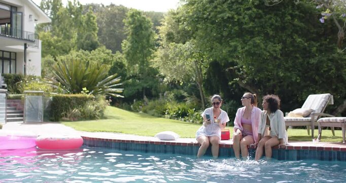 Diverse female friends showing smartphone at pool, leaning in, splashing feet, reaching red cooler