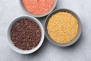 Three bowls filled with different types of lentils including red, yellow, and brown varieties arranged on a gray surface for culinary presentation or food photography