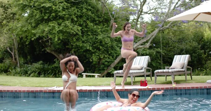 Diverse women in swimwear running, jumping into backyard pool, playing, soaking her on pink float