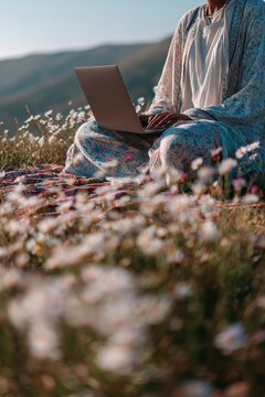 Digital nomad girl sitting cross-legged on blanket, laptop in lap, surrounded by wildflowers and hills. Springtime work-from-nature scene full of light and calm.