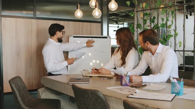 Three people collaborate in a shared workspace, focusing on a presentation while sitting at a table with paperwork and a whiteboard nearby.
