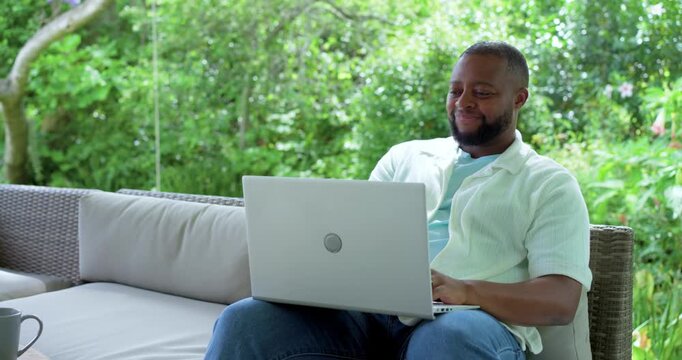 African American man wearing mint shirt on covered patio sofa, placing laptop and typing remotely