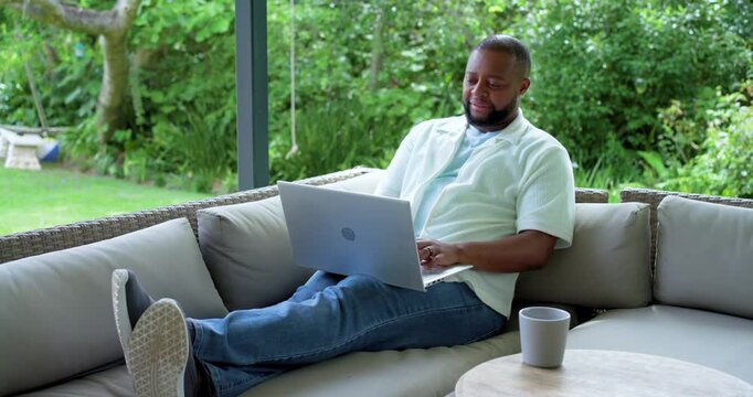 Adult African man reclining at veranda, typing reply on silver laptop, laughing at funny message