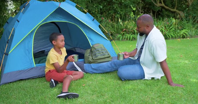 African father and son talking on grass outside blue tent, boy lifting mug, clinking mugs, bonding