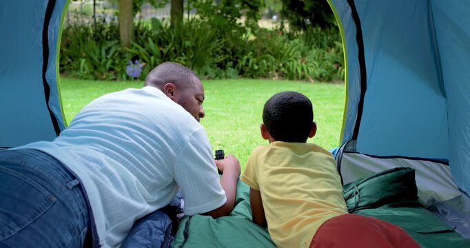 African American dad and son in pop-up tent on lawn, dad offering binoculars guiding child learning