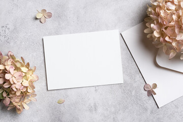 Blank invitation or greeting card mockup on a textured gray surface with dried hydrangea flowers and an envelope, featuring ample copy space for customization