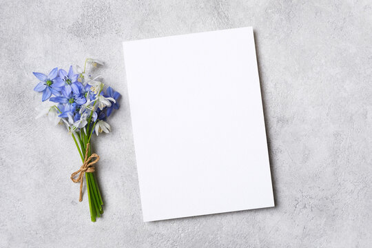 Blank invitation or greeting card mockup placed on a textured gray surface beside a small bouquet of blue flowers tied with twine for design purposes