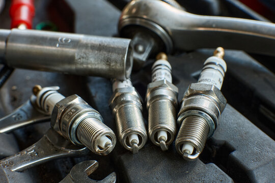 Closeup of four spark plugs with visible brand labels (NGK, Denso), resting on black engine cover beside tools, technical photography
