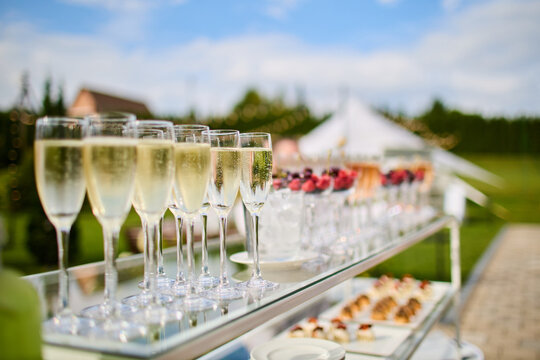 Rows of champagne flutes filled with sparkling wine on glass serving cart, outdoor wedding reception with tent in background
