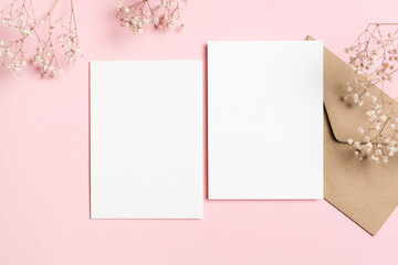 Two vertical white blank card mockups with kraft envelope and baby breath flowers on pastel pink background, perfect for wedding stationery design, flat lay