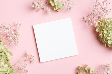 Square blank card placed on a pastel pink surface, surrounded by green hydrangeas and baby breath flowers for romantic and minimal wedding invitation mockup