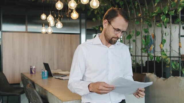 Man reads documents in a modern office with green plants and hanging lights during work hours