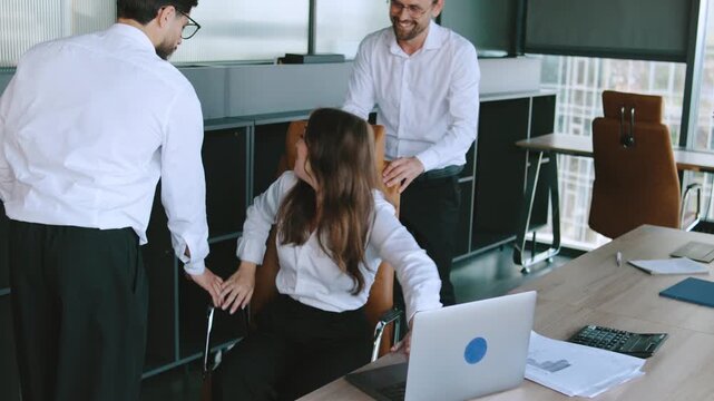 Colleagues celebrate success together in a modern office. Two coworkers express joy while one pushes a chair with a third colleague sitting in it.