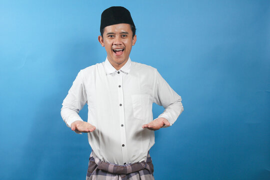 cheerful adult man wearing a traditional Muslim outfit and peci hat stands against a blue background, with his hands open as if presenting something