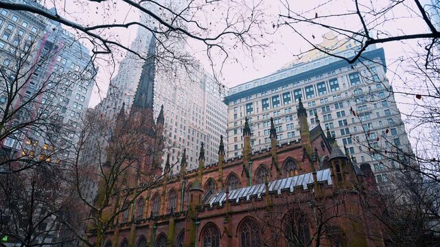 New York, USA, 2 December 2025: Spire of Trinity Church framed by bare tree branches in winter. Low angle shot looking up at the gothic spire and nearby skyscrapers through a canopy of trees.