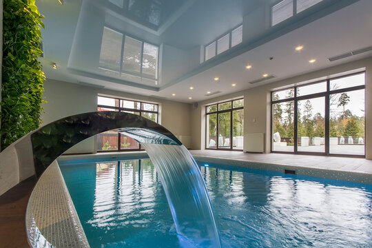 A spacious indoor swimming pool features a water fountain and large windows that let in sunlight. The room has green plants and a wooden floor
