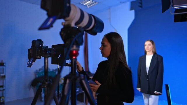 Female assistant checking video camera setup during professional studio shoot. Woman in black turtleneck stands by camera equipment with presenter in white suit in background against blue wall.