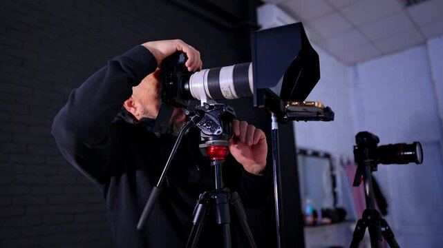 Videographer adjusting professional camera with telephoto lens on tripod. Man operates high-end digital camera with white lens in studio environment with lighting equipment and monitor.