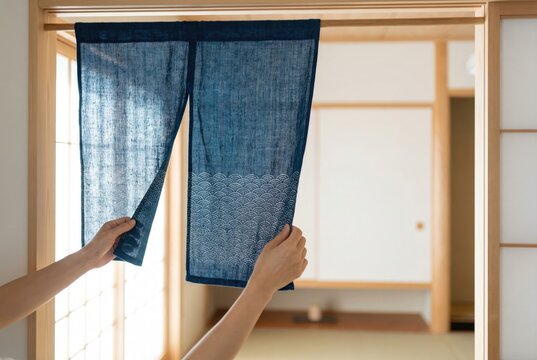 Hands hanging a traditional Japanese blue noren curtain in the doorway of a room with tatami mats.