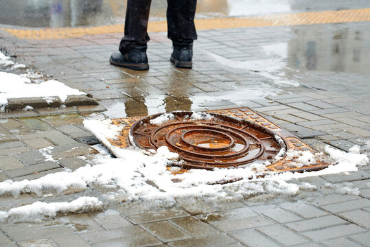 Dangerous rusted manhole cover protruding from grey brick sidewalk in the city during winter snow removal work, creating serious tripping hazard for pedestrians near slushy snow and deep puddles