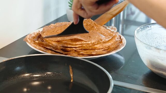Chef uses spatulas to lift a cooked crepe from hot pan and place it onto a stack of pancakes, showing homemade breakfast preparation in real time